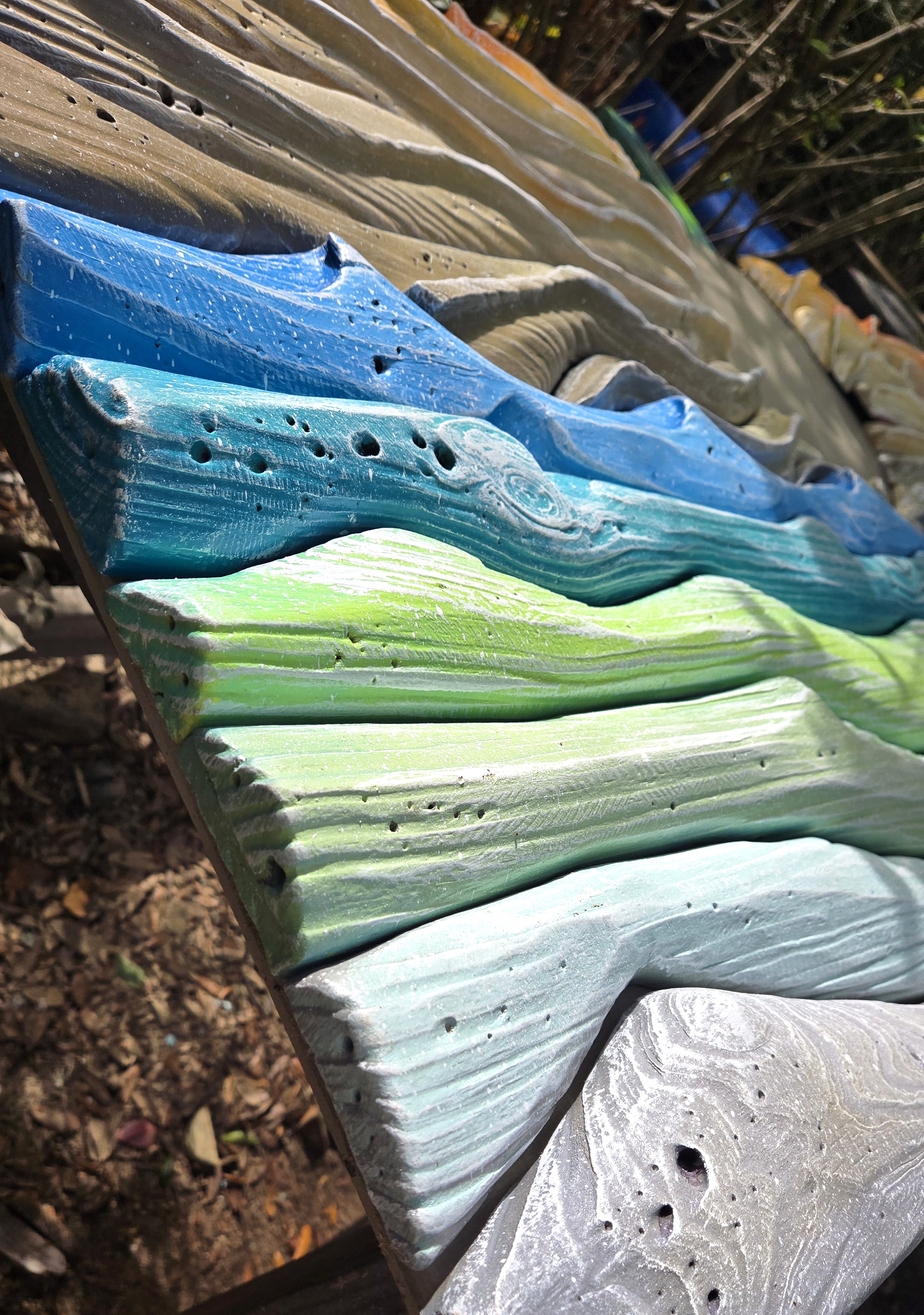 A close-up of a handcrafted wooden art piece painted with horizontal waves in shades of blue, green, and light gray, showing textured wood grain and small holes.