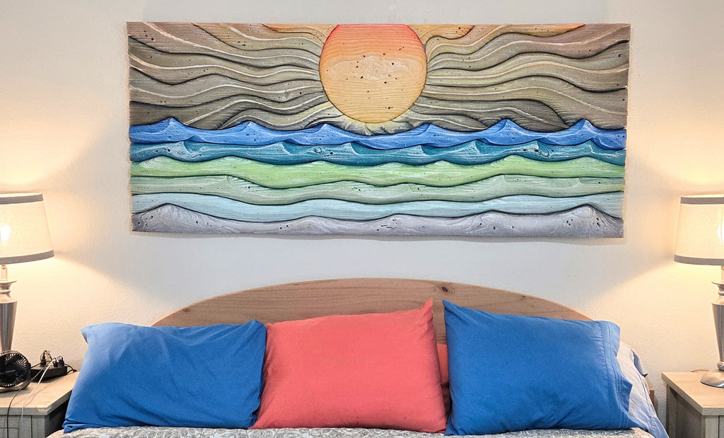 A rectangular original hand carved wall art piece by Thomas Walsh featuring layered wavy lines in earthy brown, blue, green, and gray, with a large orange sun near the top center, displayed above a bed with blue and coral pillows in a bedroom setting.