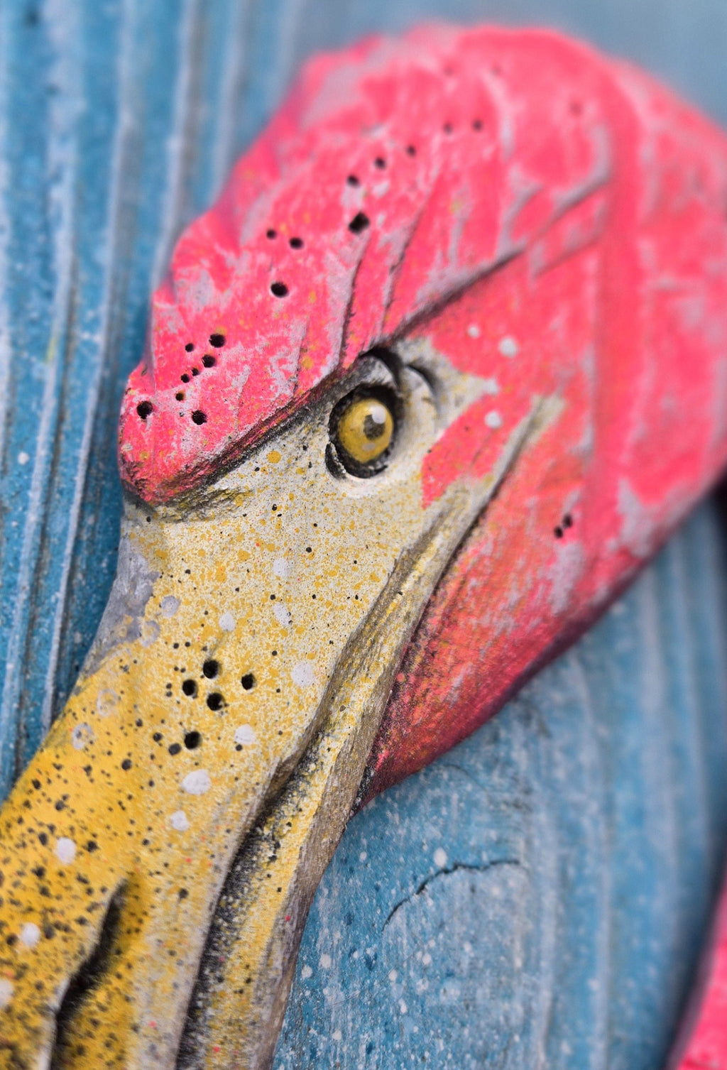 A close-up of a decorative flamingo head sculpture painted in bright pink and yellow, mounted against a weathered blue wooden background.