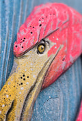 A close-up of a decorative flamingo head sculpture painted in bright pink and yellow, mounted against a weathered blue wooden background.