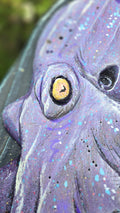 Extreme close-up of a handcrafted  Octopus face with a large yellow eye, dark pupil, and textured purple surface with paint splatters.