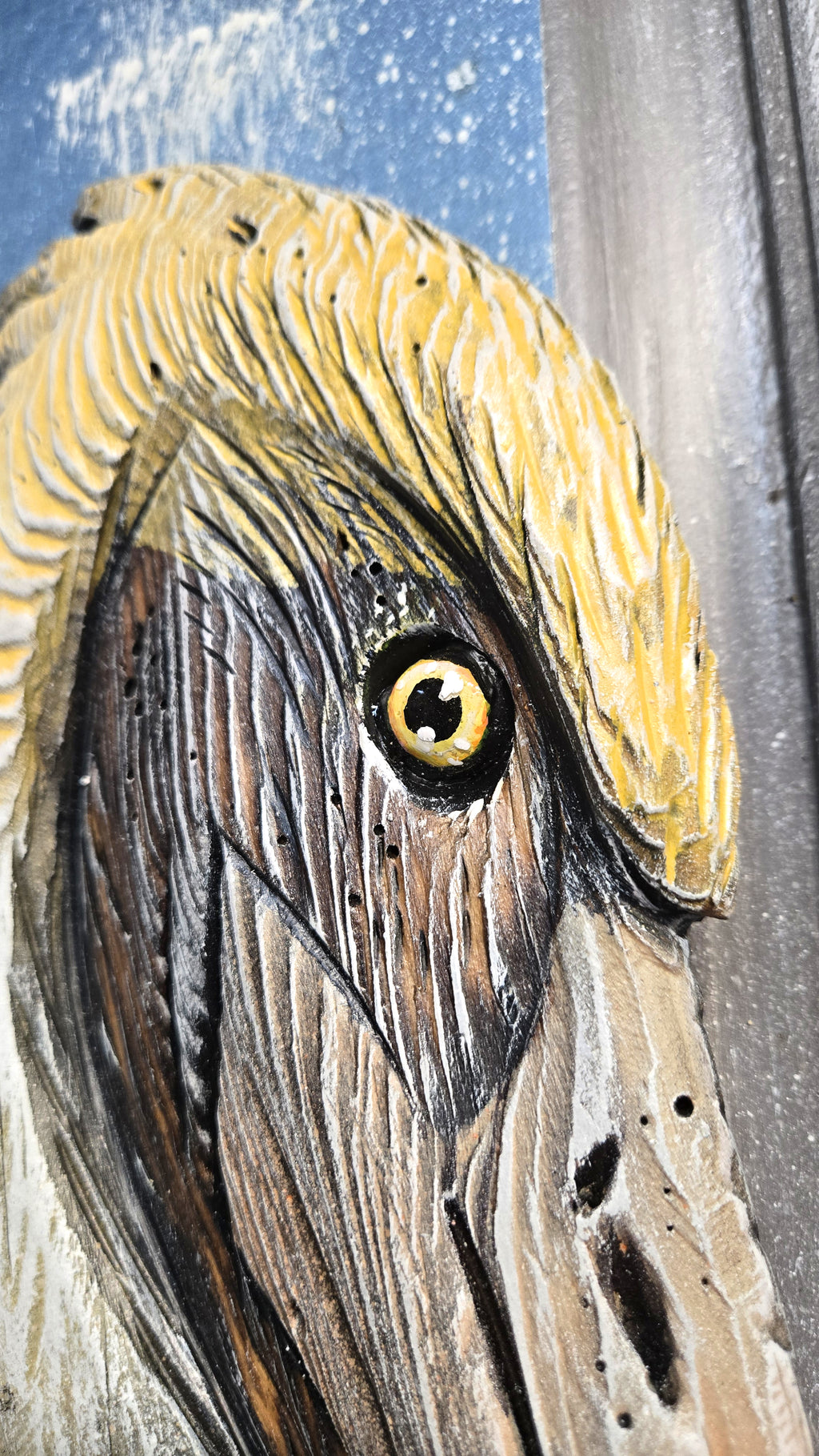 Close-up of a handcrafted carved wooden sculpture depicting a Pelican head, showing detailed wood grain, yellow feathers, and a realistic eye on a weathered blue and gray background.