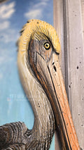 Close-up of a carved wooden pelican head with a yellow crest, brown and gray textured feathers, and a long orange beak, set against a blue sky and white cloud background. Handmade, one-of-a-kind from locally reclaimed materials Thomas Walsh at FWB Artists.