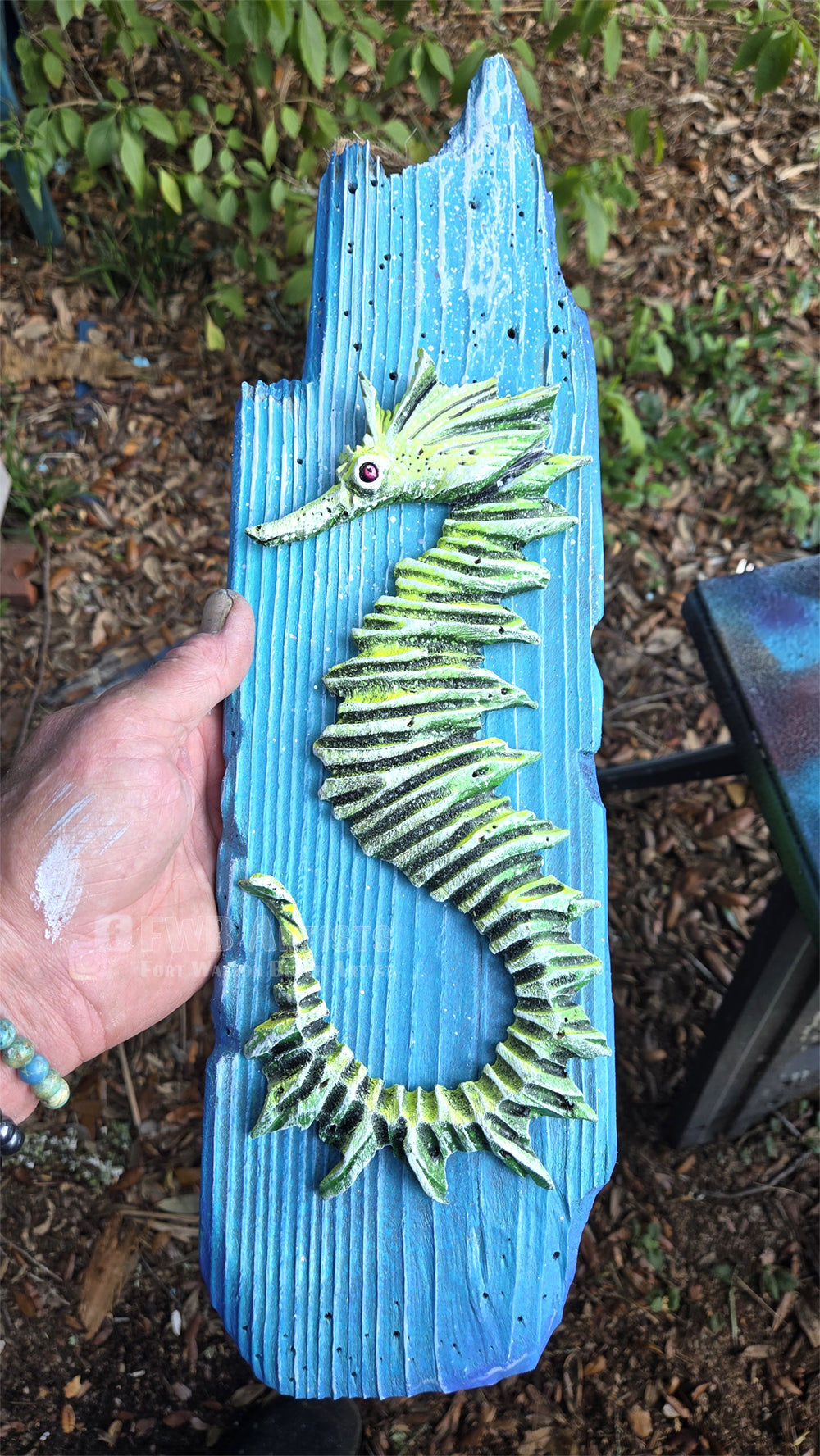 Artist's hand holds a hand carved, hand-painted wooden wall art piece featuring a green seahorse on a weathered light blue background. Handmade, one-of-a-kind up-cycled Eco-Art from locally reclaimed materials Thomas Walsh at FWB Artists.