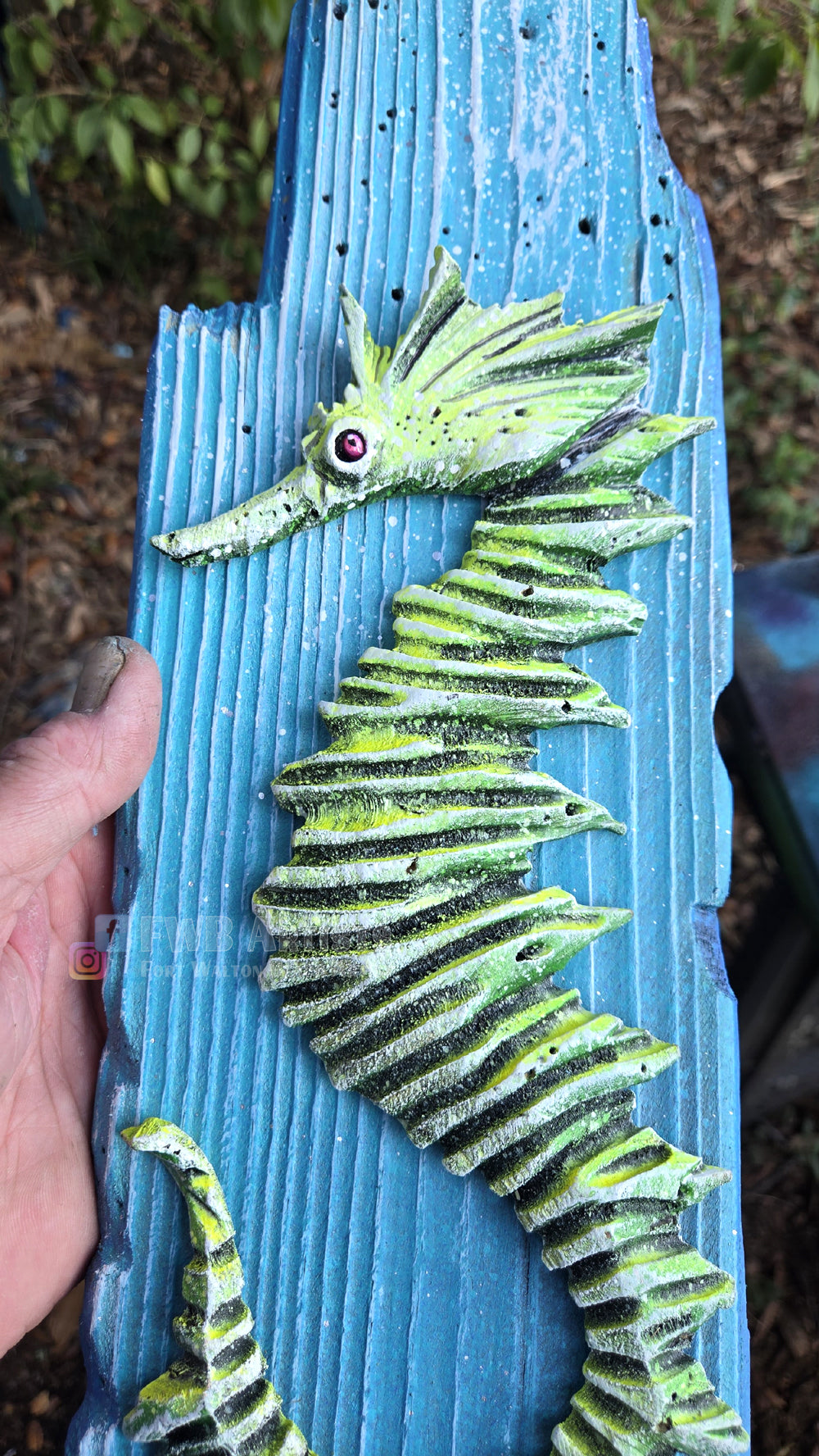 Artist's hand holds a hand carved, hand-painted wooden wall art piece featuring a green seahorse on a weathered light blue background. Handmade, one-of-a-kind up-cycled Eco-Art from locally reclaimed materials Thomas Walsh at FWB Artists.