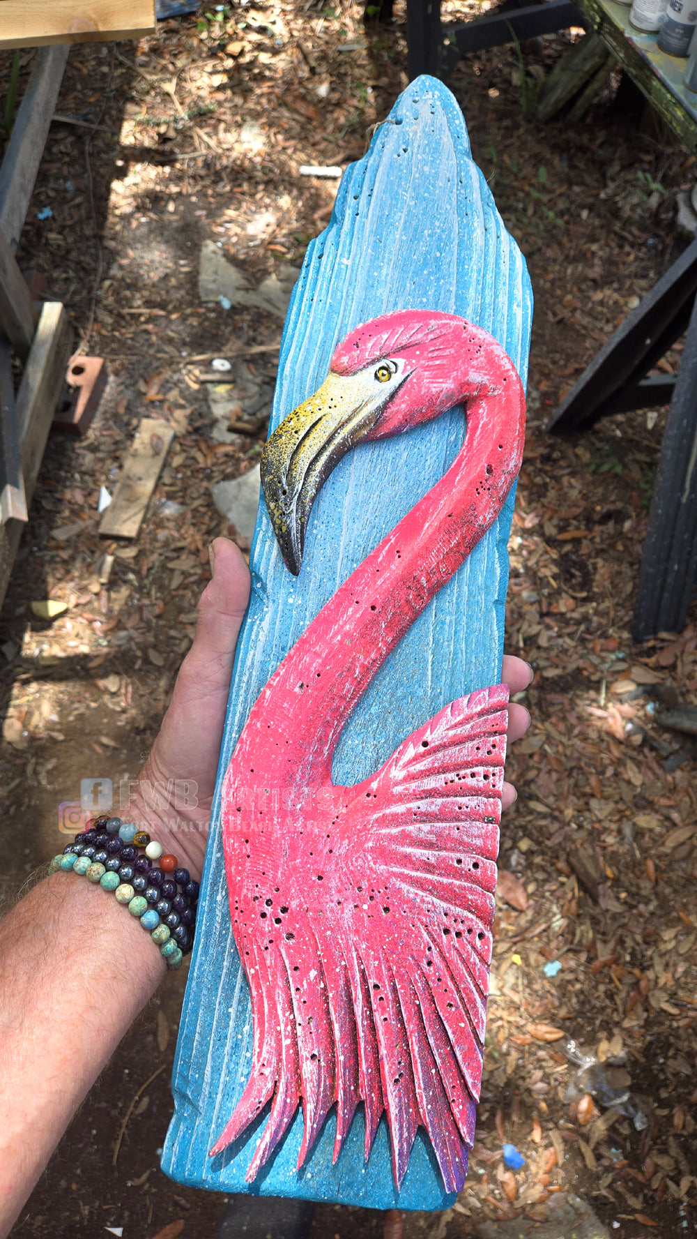 Artist's hand holds a hand carved, hand-painted wooden wall art piece featuring a bright pink flamingo on a weathered light blue background. Handmade, one-of-a-kind up-cycled Eco-Art from locally reclaimed materials Thomas Walsh at FWB Artists.