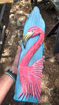 Artist's hand holds a hand carved, hand-painted wooden wall art piece featuring a bright pink flamingo on a weathered light blue background. Handmade, one-of-a-kind up-cycled Eco-Art from locally reclaimed materials Thomas Walsh at FWB Artists.
