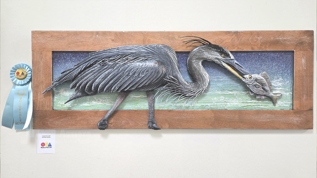 A framed wall art piece showing a detailed heron sculpture holding a fish, mounted on a wooden frame against a light wall. A blue ribbon award and a small card are attached to the left side.
