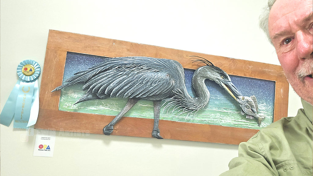 A framed 3D relief sculpture of a heron holding a fish, mounted on a wall next to a blue ribbon award and a small certificate. The artist is partially visible on the right side of the frame.