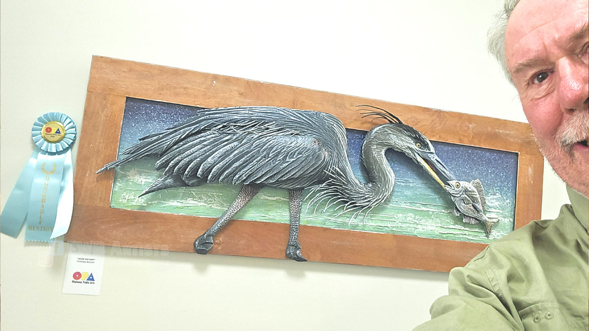 A framed 3D relief sculpture of a heron holding a fish, mounted on a wall next to a blue ribbon award and a small certificate. The artist is partially visible on the right side of the frame.