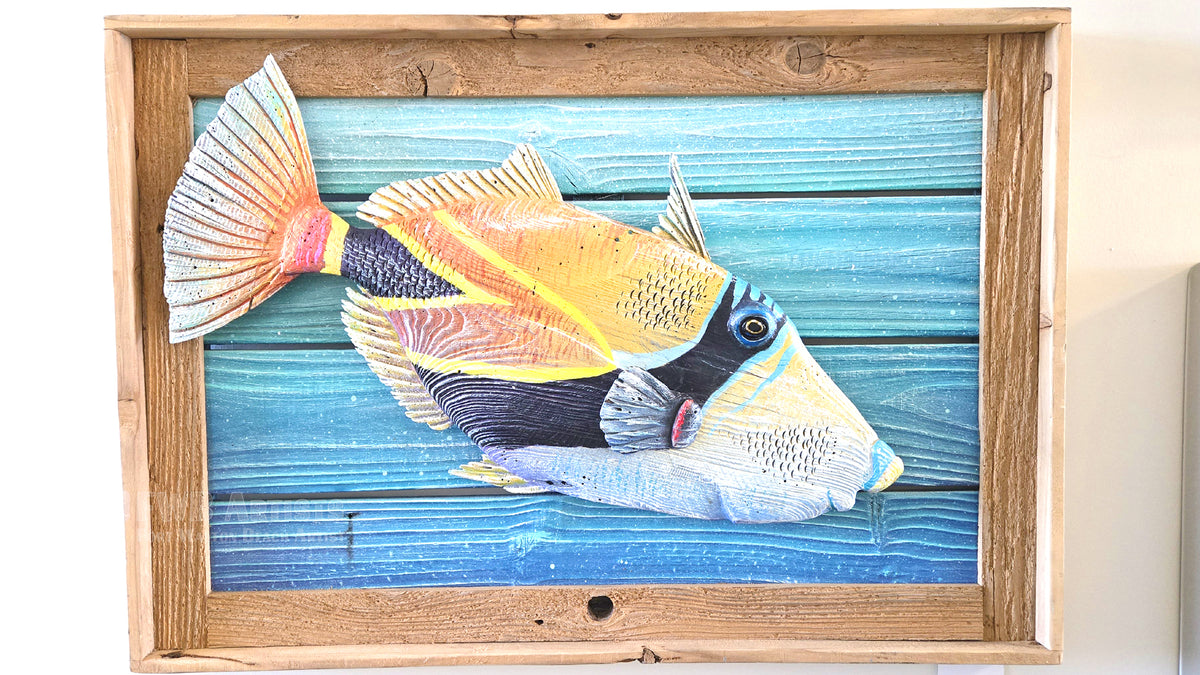 A framed wall art piece featuring a colorful hand carved and painted tropical fish on a blue wood-plank background, enclosed in a natural wood frame and photographed against a light wall.