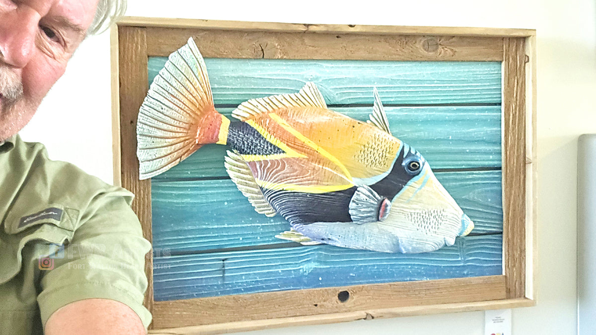 The artist poses beside a framed wall art piece featuring a colorful painted tropical fish on a blue plank background, shown indoors with part of a person visible on the left side.