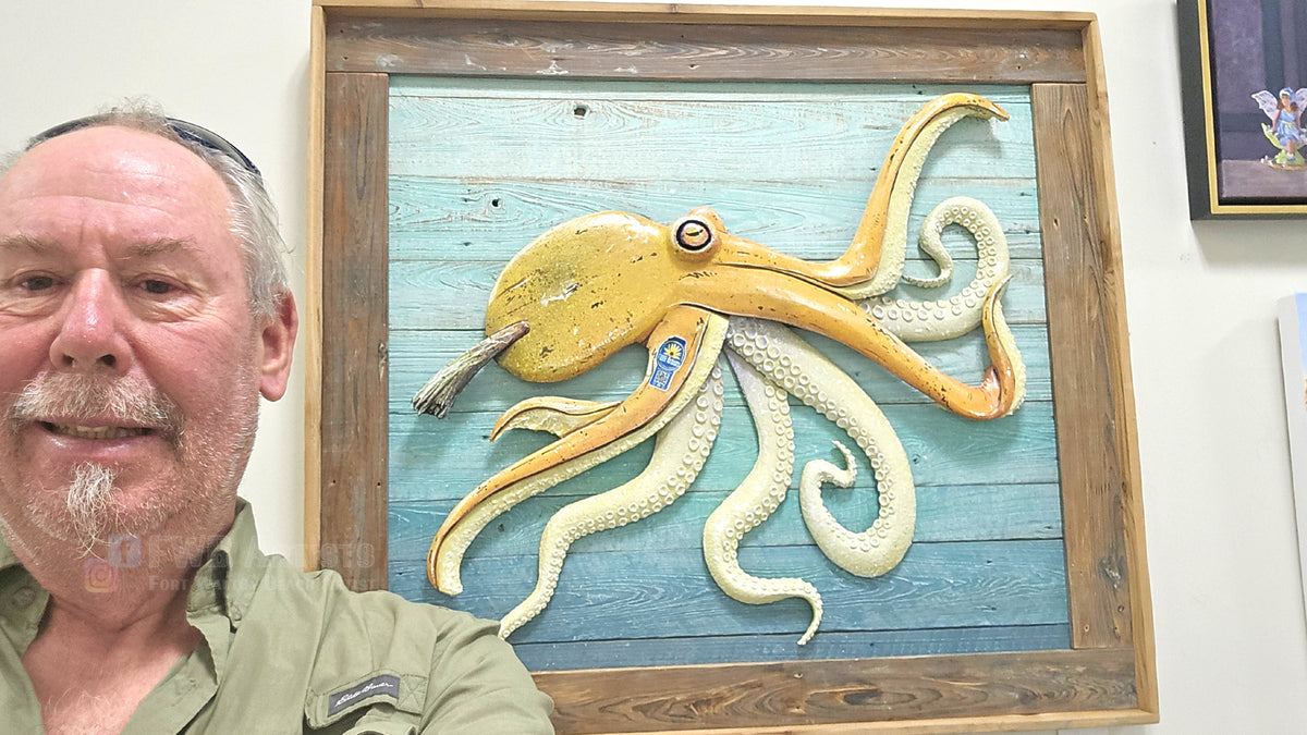 The artist smiling in the foreground next to a large framed wooden octopus wall sculpture. The octopus is painted in yellow and banana cream colors and mounted on a rustic blue wood plank background inside a dark wood frame.