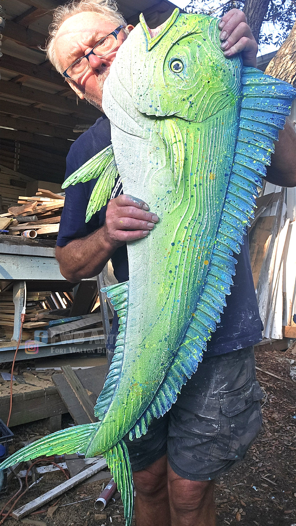 The artist at his workshop holds a large, detailed sculpture of a green and blue mahi-mahi fish.