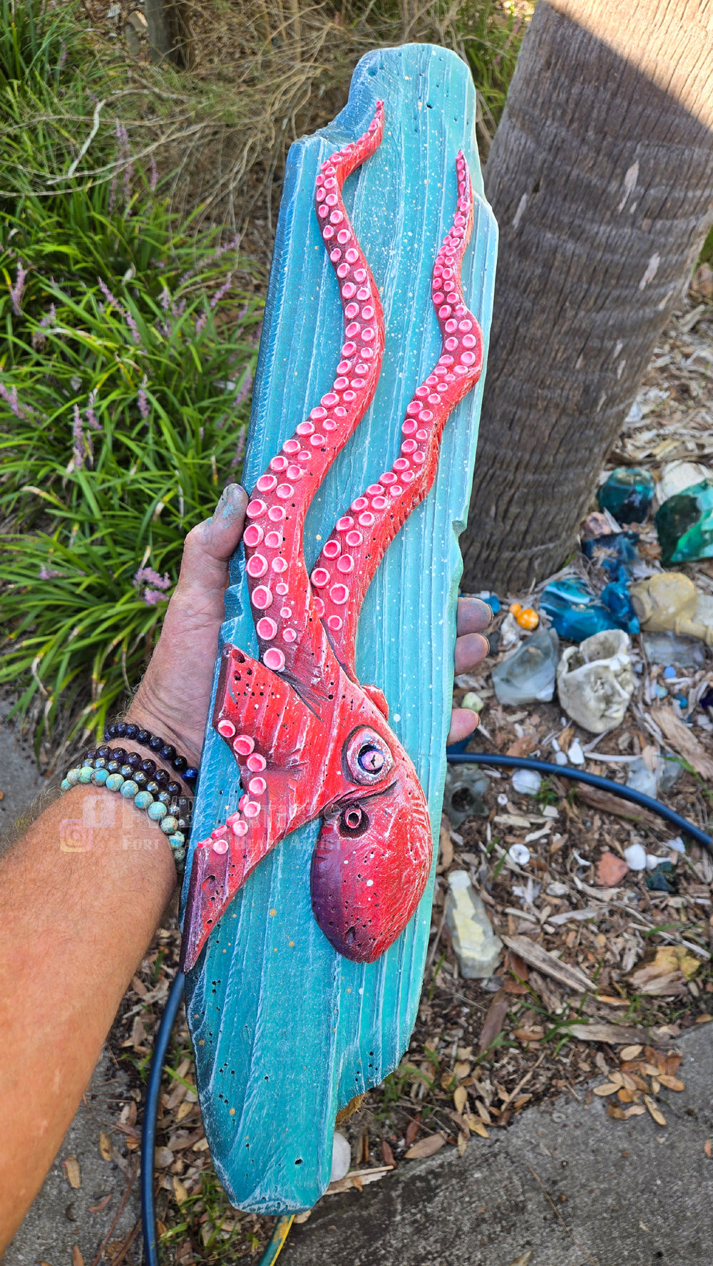 Artist's hand holds a hand carved, hand-painted wooden wall art piece featuring a red octopus on a weathered blue background. Handmade, one-of-a-kind up-cycled Eco-Art from locally reclaimed materials Thomas Walsh at FWB Artists.