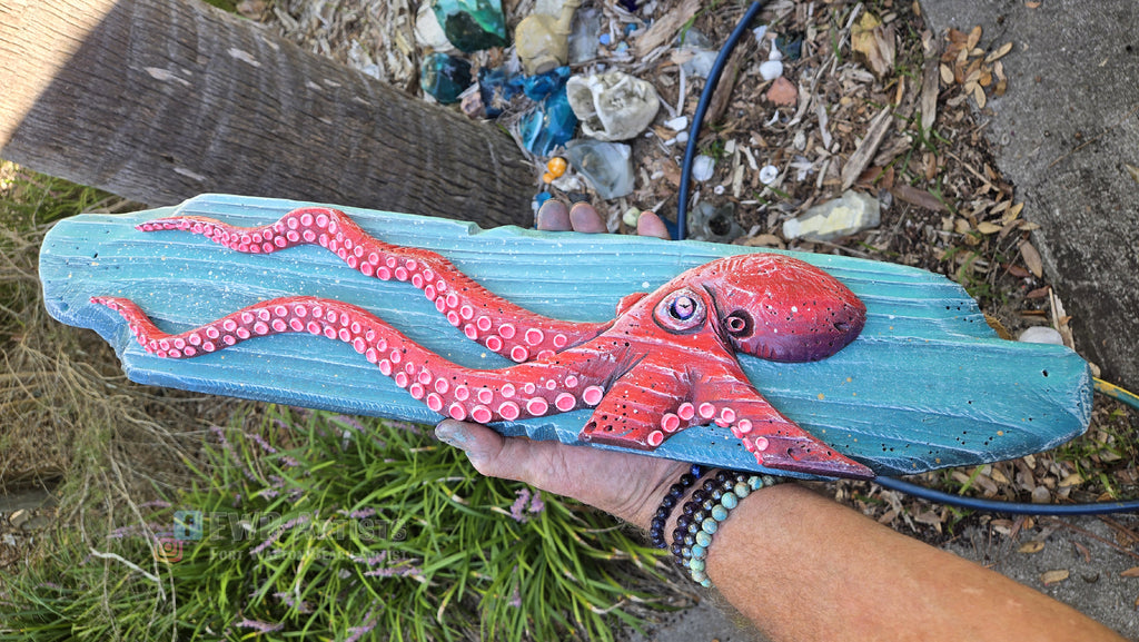Artist's hand holds a hand carved, hand-painted wooden wall art piece featuring a red octopus on a weathered blue background. Handmade, one-of-a-kind up-cycled Eco-Art from locally reclaimed materials Thomas Walsh at FWB Artists.