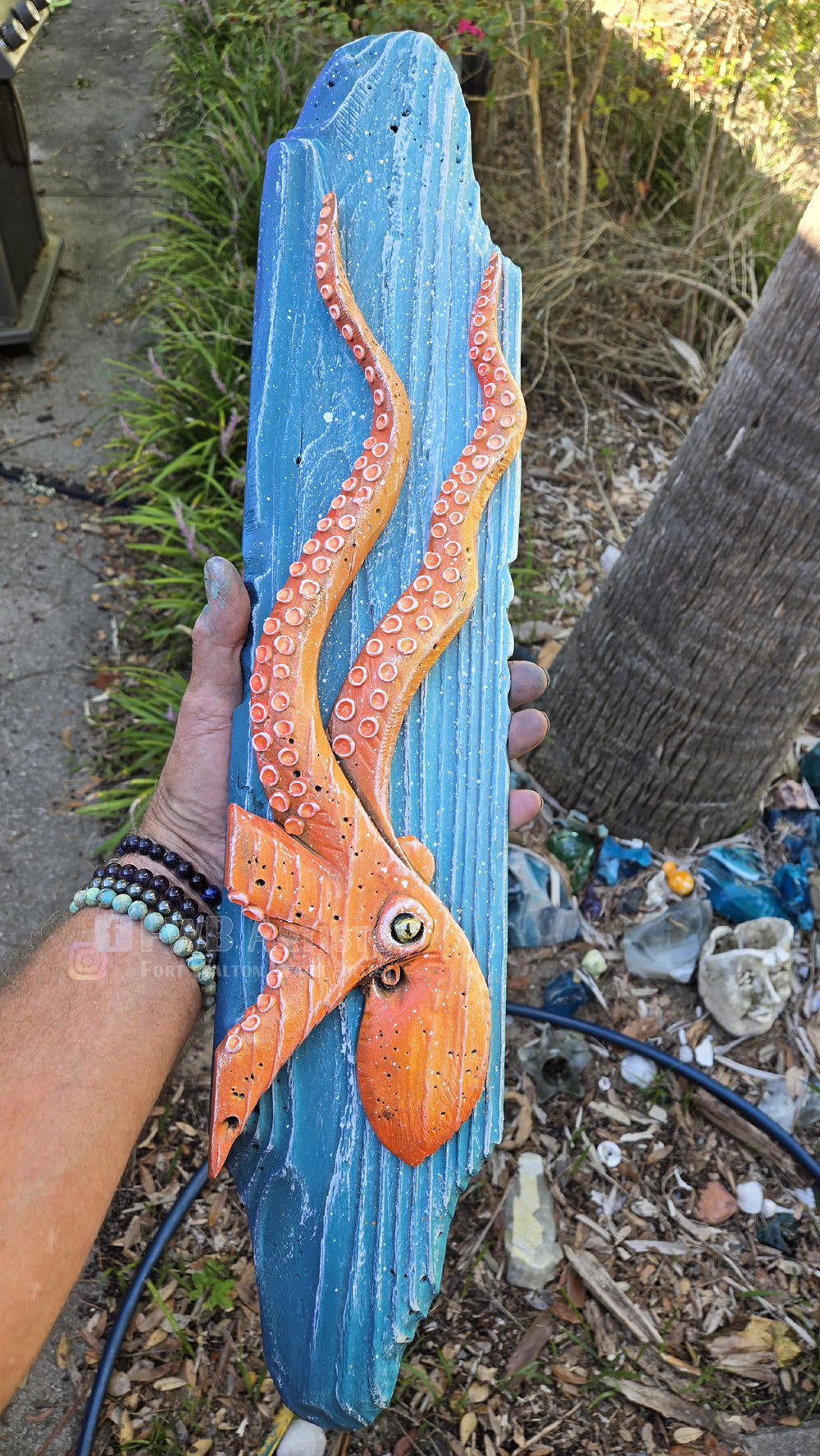 Artist's hand holds a hand carved, hand-painted wooden wall art piece featuring a orange octopus on a weathered blue background. Handmade, one-of-a-kind up-cycled Eco-Art from locally reclaimed materials Thomas Walsh at FWB Artists.