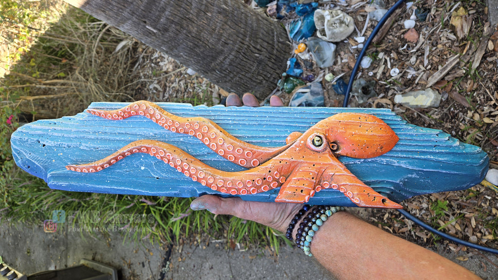 Artist's hand holds a hand carved, hand-painted wooden wall art piece featuring a orange octopus on a weathered blue background. Handmade, one-of-a-kind up-cycled Eco-Art from locally reclaimed materials Thomas Walsh at FWB Artists.