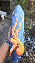 Artist's hand holds a hand carved, hand-painted wooden wall art piece featuring a Flo-orange octopus on a weathered blue background. Handmade, one-of-a-kind up-cycled Eco-Art from locally reclaimed materials Thomas Walsh at FWB Artists.