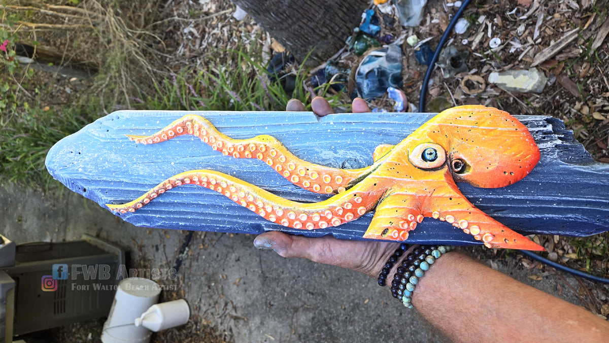Artist's hand holds a hand carved, hand-painted wooden wall art piece featuring a flo-orange octopus on a weathered blue background. Handmade, one-of-a-kind up-cycled Eco-Art from locally reclaimed materials Thomas Walsh at FWB Artists.