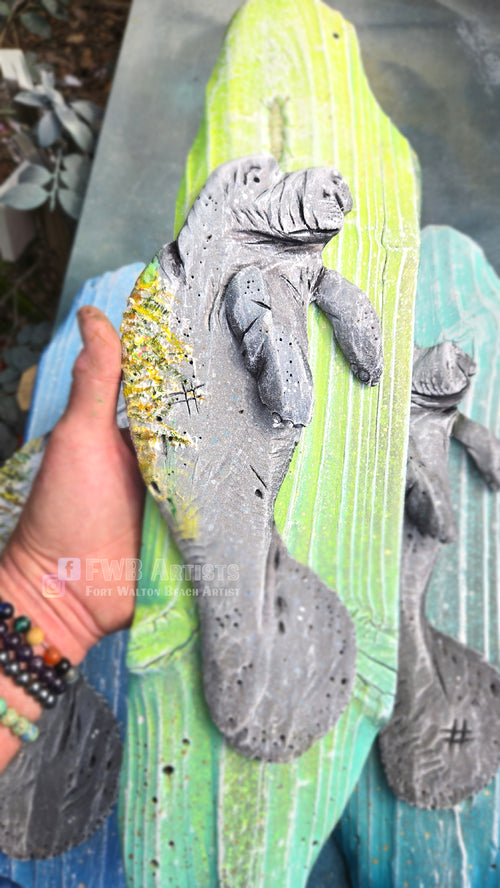 Artist's hand holds a hand carved, hand-painted wooden wall art piece featuring a gray baby manatee on a weathered light green background.