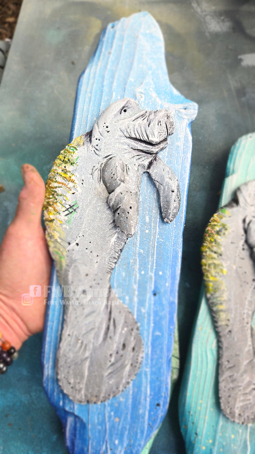Artist's hand holds a hand carved, hand-painted wooden wall art piece featuring a gray baby manatee on a weathered light blue background.