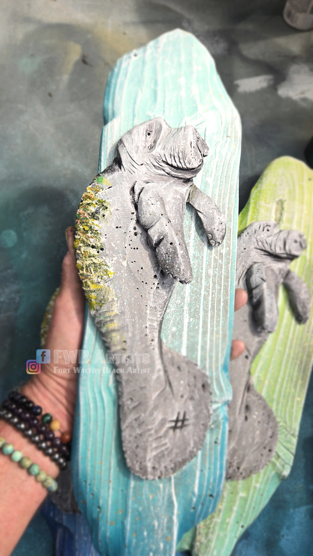 Artist's hand holds a hand carved, hand-painted wooden wall art piece featuring a gray baby manatee on a weathered light teal background.